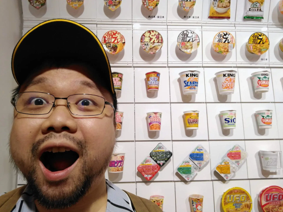 A man standing in front of a wall covered in dozens of containers of various types of instant ramen and udon noodles. The man's facial expression shows amusement.