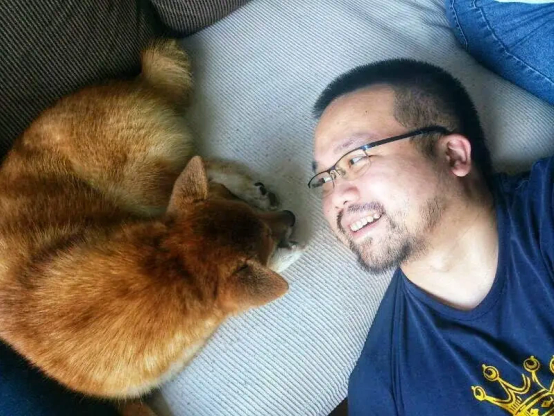 A top-down shot of a man lying down, looking into the eyes of a shiba inu dog. The dog has curled up into a resting position.