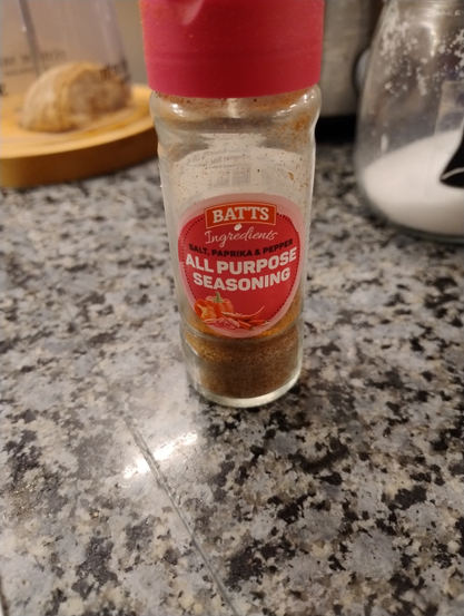 Container of Batts All Purpose Seasoning with about 1/3 to  1/4 contents left. It is on a kitchen counter with cheese and salt in the background.