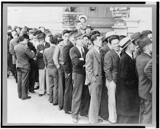 A black and white photograph depicting a group of individuals, presumably Japanese Americans or those of Japanese ancestry during World War II era. The people are seen lined up in an orderly fashion at what appears to be the Civil Control Station for registration as per Army's exclusion order No. 20.

They appear to wear formal attire such as suits and hats typical of men from that period, with some wearing ties or glasses which is indicative of middle-class individuals during the era. One person stands out in this line holding a child on their shoulders while others seem waiting patiently for their turn at registration. The setting seems urbanized given what appears to be buildings behind them.

The photograph suggests a historical context likely taken within San Francisco, California around April 1942 as mentioned in caption information provided which also references the War Relocation Authority centers established during World War II that housed evacuees of Japanese ancestry throughout their duration. The expressions on these individuals' faces vary from somber to engaged with one another or lost in thought.

This image is part of a collection by photographer Dorothea Lange, known for her work capturing the social issues and human emotions around wartime events during World War II, particularly relating to internment camps across America. This specific photograph highlights not just an administrative procedure but als [...]