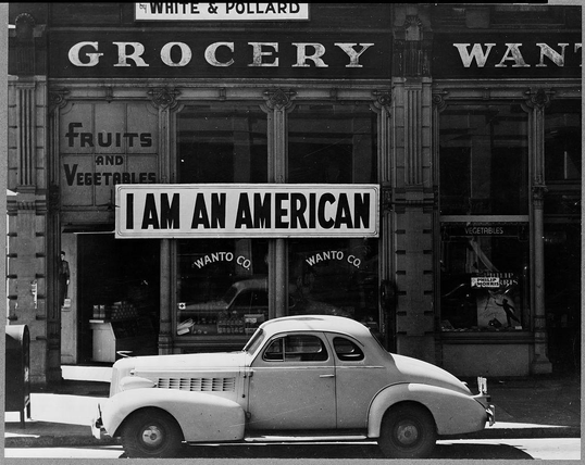 The image is a black and white photograph depicting an old-fashioned store with signage that reads "Grocery" above the entrance, indicating it's likely a grocery shop. The storefront displays various goods such as fruits and vegetables, suggesting its purpose in providing food items to customers.

A prominent feature of this photo is a large sign placed on the window reading "I AM AN AMERICAN," which suggests an expression or statement being made by someone associated with the store, possibly reflecting feelings about identity during that historical period. The context provided indicates this image was taken during World War II in Oakland, California, specifically at 401-403 Eighth and Franklin streets on December 8th.

The vehicle parked outside is an older model car typical of mid-century design with rounded edges and a shiny surface reflecting the environment around it. It adds to the historical context of the image by indicating its time period before modern automotive styles became prevalent.

Overall, this photograph captures a moment in American history during World War II when personal identity was being questioned due to racial tensions as seen through the lens of an individual's assertion of their nationality amidst wartime evacuations and restrictions on people of Japanese descent.