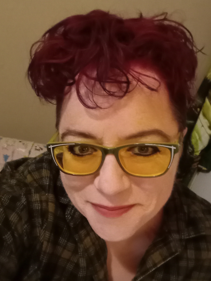 Vayl Larkin is pictured, a nonbinary transmasculine person with fuschia hair that is curly and short. (Fuschia is a bright, bold cross between pink and purple). Their button shirt is flannel and their green glasses frame their rounded heart-shaped face. Their nose is small, as is their mouth, but their eyes look bigger as a result so it's all relative, you know? I like knowing only people reading the description are going to know that's how I feel about that. Others can just try to guess!