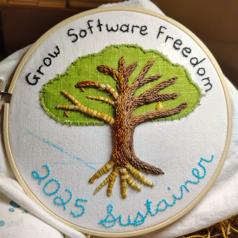 A work-in-progress picture of my embroidery. A tree with the words "Grow Software Freedom" above the canopy. The phrase "2025 Sustainer" is below the tree roots.

Brown bark is covering most of the tree branches. Only one root is covered in bark. The bare branches have stripes of yellow yarn circling them, which the bark yarn will loop into.