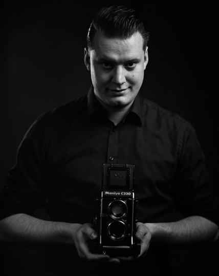 Black and white photo of white male, looking at camera and holding a Mamiya C330 TLR camera at waist level.