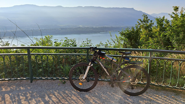 mon vélo, un VTT gris foncé, posé contre une barrière. en contrebas, le lac du Bourget sous le soleil.