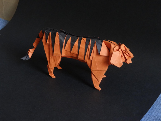 un tigre en origami, orange rayé de noir. il est posé sur une feuille noire.