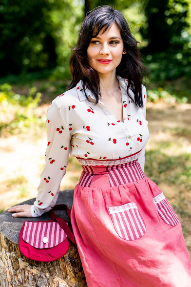 Dans une clairière ombragée, une jeune femme brune est assise sur une souche d'arbre. 
Elle porte un chemisier blanc à motifs de cerises, et je lui ai réalisé une jupe corsetée et un sac assorti. 
La jupe est en lin rose, munie de 2 poches plaquées sur le devant en coton rayé rose et rouge, et surmontées de dentelle crème. 
La partie corsetée de la jupe qui permet de marquer la taille est réalisée en algorithme avec les tissus en lin uni et le coton rayé. Elle est aussi surmontée d'une dentelle crème. 
Le petit sac de forme arrondie est réalisée en coton rouge framboise, et son rabat est réalisé avec le coton rayé. 

Modèle : Julie 
Photographe : Namidaël Photographies 