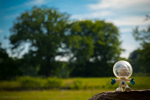 The image shows a small Smurf astronaut toy standing on a wooden surface in a field. In the background, there are large trees and a blue sky with some clouds. The astronaut has a bubble-like helmet and is wearing a white spacesuit with blue gloves. It looks like the toy is enjoying a sunny day in the countryside.