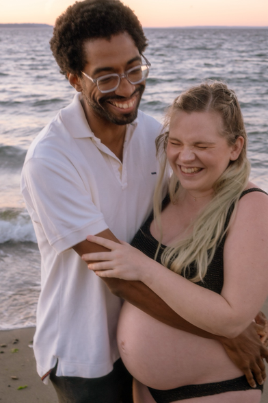 A Black man holds a pregnant, white nonbinary person in front of the ocean. They are both smiling and the nonbinary person has their eyes closed because they are laughing so hard.