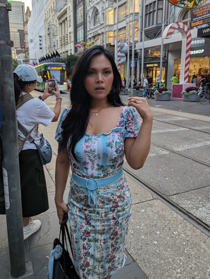 A woman stands near the tram tracks. She is wearing blue dress. Shes in a middle of a sneeze