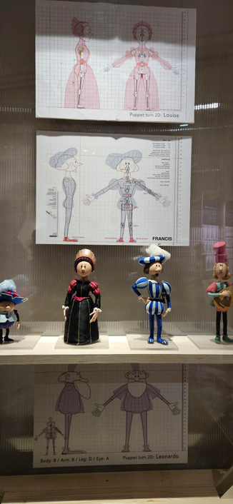 4 puppets in a row at an exhibition. Those puppets are from the movie Inventor, dressed in Renaissance attires. From the left to the right there is a small and round cartoony boy, with a big feather hat, a black haired woman with a crown and a black dress, a man with a moustache and blue striped clothes and a blond haired man with a luth and pink and green outfit.