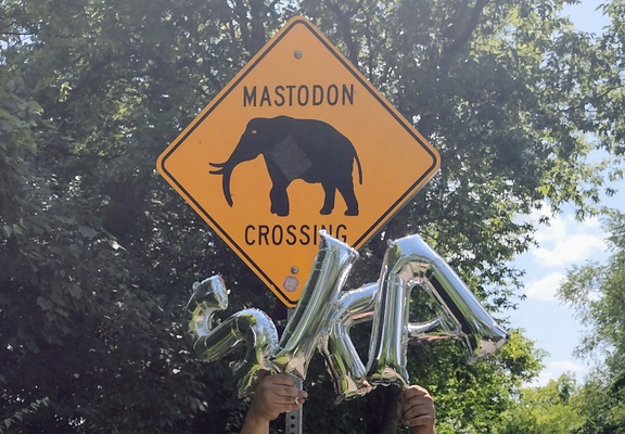 A yellow diamond "Mastodon Crossing" sign. And underneath the sign are three silver balloons spelling the word "SKA."