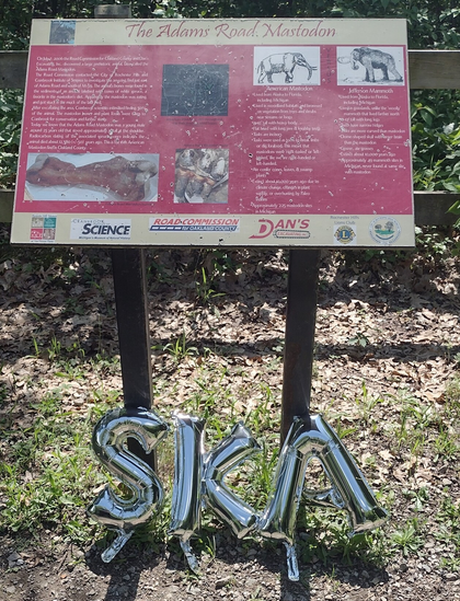 A placard for the Adams Road Mastodon. Underneath are three silver balloons spelling the word "SKA."