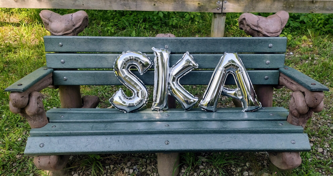 A park bench partially of replica mastodon bones. Atop are three silver balloons spelling the word "SKA."