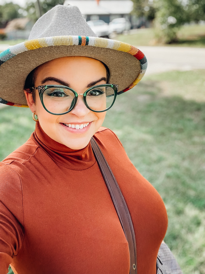 Indigenous woman smiling in beaded hat, green glasses, while wearing a leather crossbody bag.