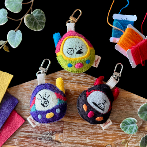 A product picture of three felt tamagotchi-like toys. The top one is pink and green with embroidered buttons and a blue antenna. The creature embroidered on the screen is a sad blob next to a stinky poop. The bottom left one is purple, red and green with grey and yellow buttons and a yellow antenna. Embroidered on it is an egg thats’’s currently hatching. The bottom right one Is black and red with red devil horns and orange and red buttons. On the screen is embroidered a tombstone.
All models have an ivory cotton ribbon with a gold or silver lobster clasp. 