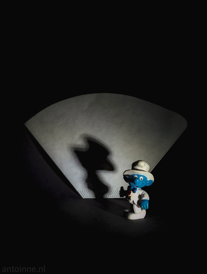 A small blue toy Smurf dressed as a ringmaster standing in front of an oldskool coffee filter. The lighting casts a large shadow onto the filter. The figurine is wearing a white suite and hat. The background is dark black. 