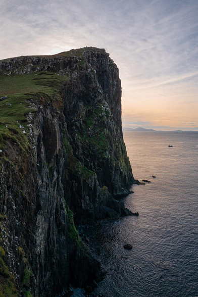 Large cliffside overlooking the ocean with some mountains in the far background. Shot is taken at sunset and has a warm glow coming from behind the cliff.