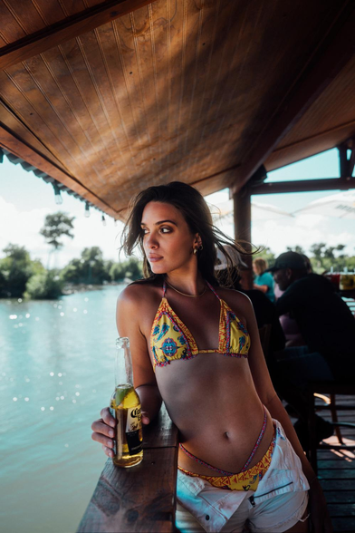 Model type beauty  in bikini holding a beer while leaning against a wooden rail overlooking a river in the tropics