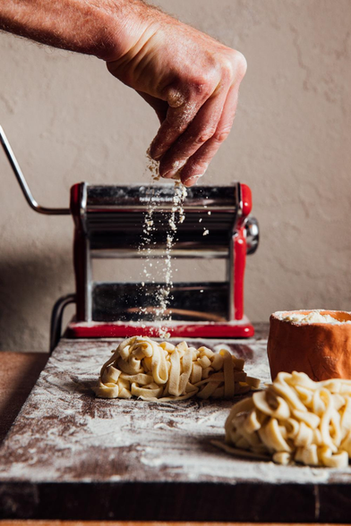 hand dropping flour over freshly made pasta. There is a past rolling machine in the background and two mounts of pasta on the board which has a good amount of flour all over. The feel is very rustic.