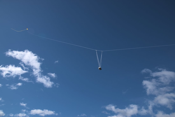 Sobre un cielo azul intenso se ve una linea blanca que la atraviesa de derecha a izquierda. En medio de la linea puede verse la cámara amarrada con varias cuerdas y al final de la línea una cometa amarilla con una larga cola.