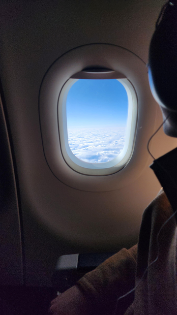 An airplane window shows a view of a bright blue sky above a vast expanse of fluffy white clouds. A person's shoulder and arm, wearing a dark brown jacket, are visible in the lower right corner, partially obscuring the view. A portion of the person's headphones is also visible. The airplane's interior is dark gray.