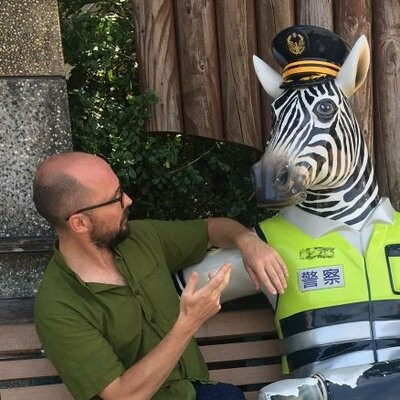 Picture of Will Buckingham, sitting on a bench in Taiwan, talking about philosophy with a life-sized sculpture of a zebra. The zebra is dressed as a Taiwanese police officer, complete with hi-vis jacket and police officer's hat.