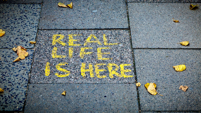 Gelbe gepinselte Großbuchstaben auf dunkelgrauem Bürgersteig. Dort steht: Real Life is here
