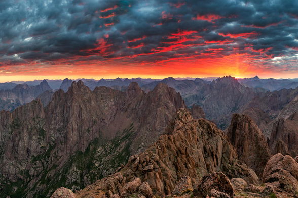 A breathtaking sunrise from 14,000 feet in Colorado's San Juan Mountains. Jagged peaks stretch across the horizon under dramatic red and orange clouds, showcasing the rugged beauty of this alpine landscape.