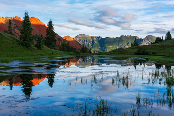 A serene high mountain lake near Silverton, Colorado, reflects the vivid red hues of sunlit peaks and the surrounding evergreen trees. The calm water mirrors a clear blue sky with soft clouds, creating a tranquil alpine scene.