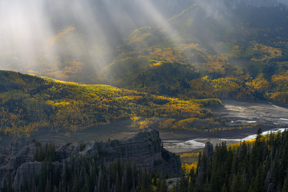 Golden aspens glow in the fall light, illuminated by sunbeams breaking through the clouds over Ridgway, Colorado. A rugged landscape of pine trees and dry riverbeds stretches across the valley, bathed in warm, radiant light.