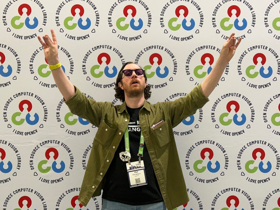 A man wearing a green button up shirt (unbuttoned) with a black t-shirt underneath. He is gesturing in front of a backdrop with the OpenCV logo repeated in a staggered pattern, giving the vague impression of a cult leader.