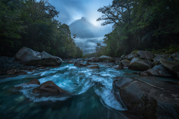 an image taken from the middle of a New Zealand stream looking through rocks & trees towards a mountain in the distance with light shining through