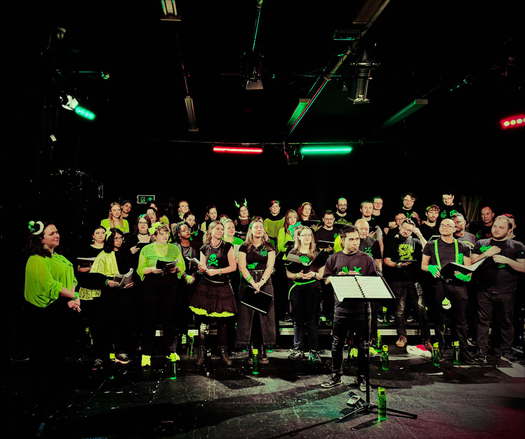 A group of singers dressed in neon green and black, on stage at a concert (it's Ready Singer One!) - one chorister has stepped up to the lectern to introduce a song. Most have a logo on their t-shirt with something nerdy. 