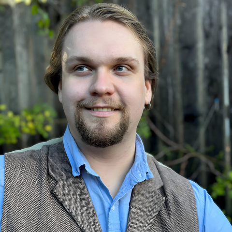 A blond, blue-eyed man in a blue-collar shirt and herringbone vest looks into the distance and smiles.