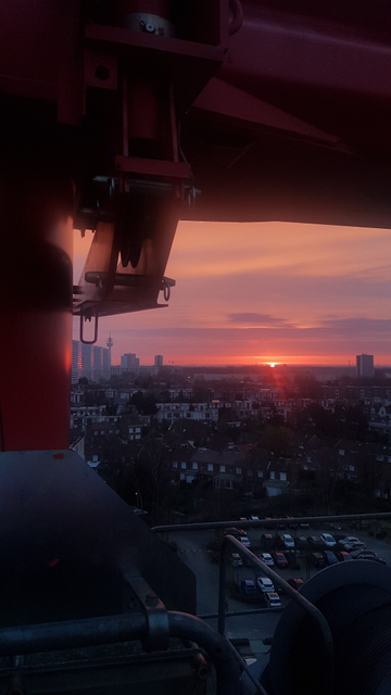 lever de soleil pourpre, rose et violet, vue du dessous d'une grue avec la ville en bleu et petit en bas, le soleil perce son premier rayon très rouge et des immeubles au loin reflètent la couleur du ciel