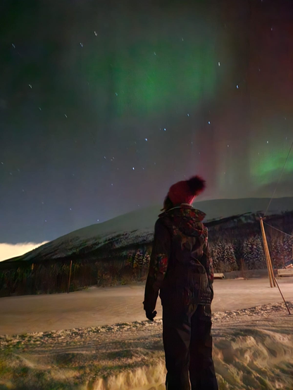 personne de dos avec un bonnet rouge a pom-pom qui regarde le ciel la nuit le sol est entièrement recouvert de neige et une montagne est non-loin recouverte d'arbres enneigés, le ciel affiche des étoiles et des aurores boréales vertes et rouge