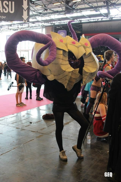 dans une foule a l'intérieur d'un très grand hangar, un costume de créature flottante avec un oeil gigantesque et trois tentacules se tient au milieu, alors que les jambes de la personne en-dessous sont visibles entièrement habillée en noir et avec une pose de pin-up