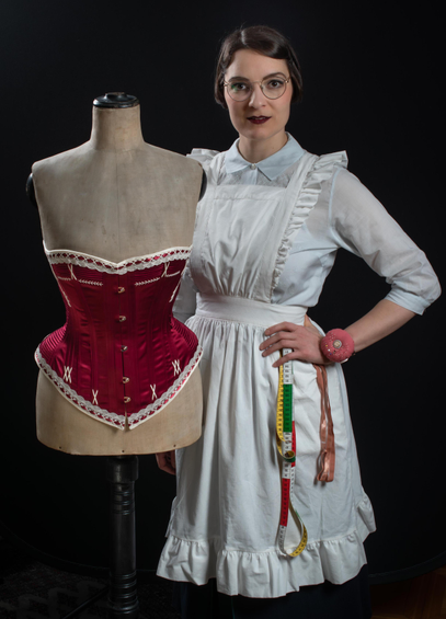 Cette photo représente la corsetière suisse Lucie Violette Vallez, dans une tenue de couturière rétro. Elle pose à côté d'un mannequin de couture antique qui est vêtu de l'une de ses créations : une reproduction historique d'un corset victorien.