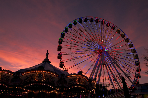 Photograph of a carnival at sunset/night, at an upward angle, showing the roofs of a carousel, a ferris wheel, and an orange, pink, and purple sky. All the amusement park equipment is lit up with string lights.