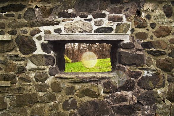 A photograph of a small window in a stone wall, looking out on a grassy expanse. There is a full moon overlaid transparently over the grass, centered in the window.