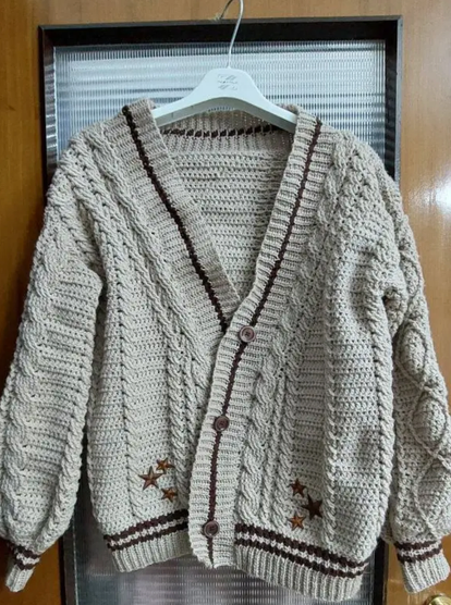 Crochet cabled cardigan inspired by Taylor Swift's Folklore cardigan. It's made out of a beige or sand brown yarn, with accents in chocolate brown yarn. It includes brown star patches on the front and three buttons. The color scheme is inspired by the album Evermore.