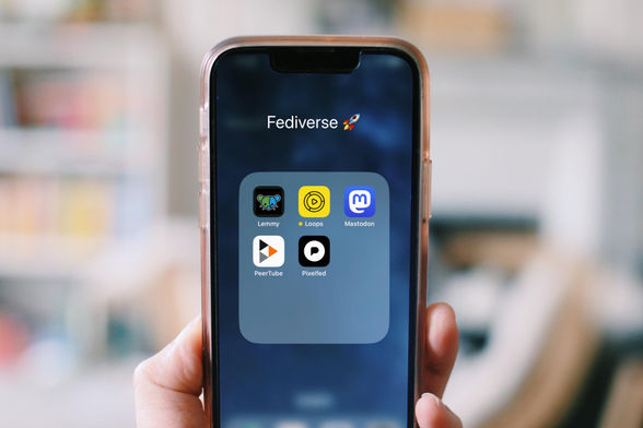 a photo of a hand holding up a smartphone. On screen you see the folder "Fediverse" and 5 app icons: Lemmy (actually a shortcut to the website), Loops, Mastodon, PeerTube and Pixelfed