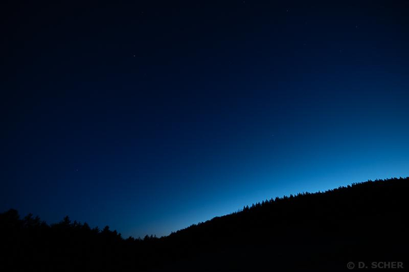 A conifer-covered ridge line emerges against the last evening light, while the upper sky is already dark and revealing the first stars.