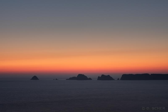 Just after sunset, the sky shows a gradation of colors from dark blue to blazing orange and red. The ocean is still slightly visible in the lower half of the photo. A string of rocky islet silhouettes lies between sea and sky across the entire width of the scene.