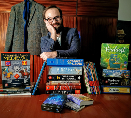 A male person with glasses and beard looking at the camera, leaning over a bunch of books and board games.