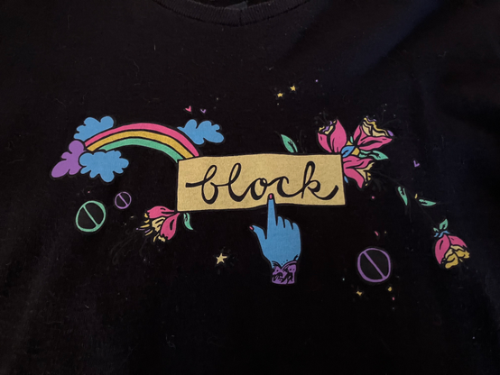 A photo of the front of a black T-shirt (although it's not obvious it's a T-shirt) which has a yellow rectangle with the wood "block" written in script. A finger on a blue hand points to the word.The rectangle is surrounded by images of flowers, plus a rainbow.