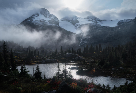 A mountain and lake scene with dappled sunlight coming through the clouds. A waterfall is coming down the mountain in the distance and the lake is surrounded by trees, some with autumn foliage.