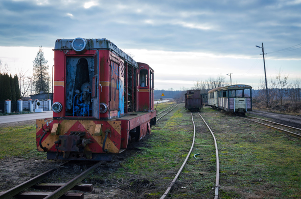 Three narrow gauge tracks next to a road in the countryside. First track contains Romanian-made FAUR L45H (PKP Class Lxd2) diesel locomotive in a terrible condition, ripped open and damaged, windows long smashed, graffiti on. Some similarly miserable wooden carriages can be seen further away in the distance