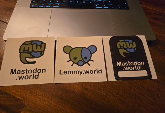 Two mastodon.world stickers and one lemmy.world sticker in front of a macbook